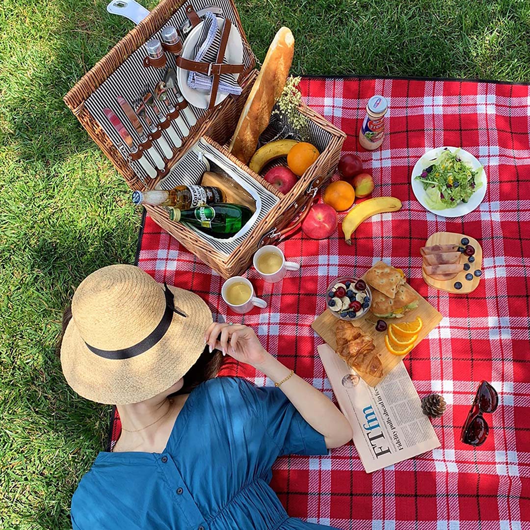 a woman lies on a picnic blanket next to a picnic basket and food.